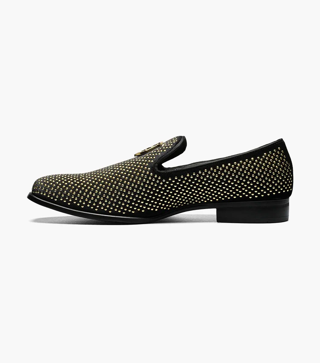 The Stacy Adams - SWAGGER Studded Slip On - Black and Gold - 25228-715 is a black loafer adorned with gold studs and a decorative emblem on top, featuring a comfortable memory foam insole.