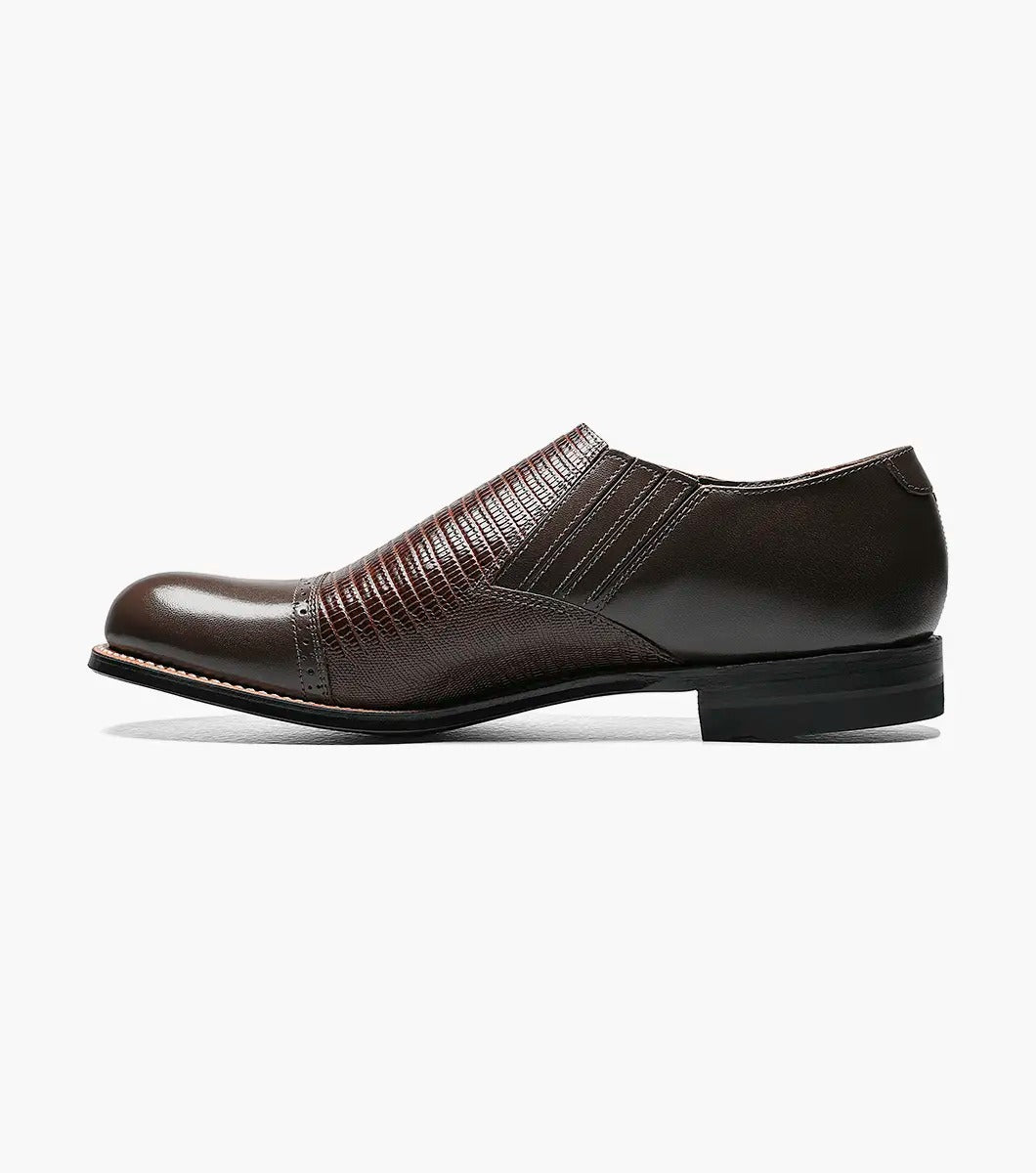 The Stacy Adams - MADISON Cap Toe Slip On in brown is shown in a side profile view against a white background. This shoe features embossed detailing and a low heel, crafted with Goodyear welt construction.