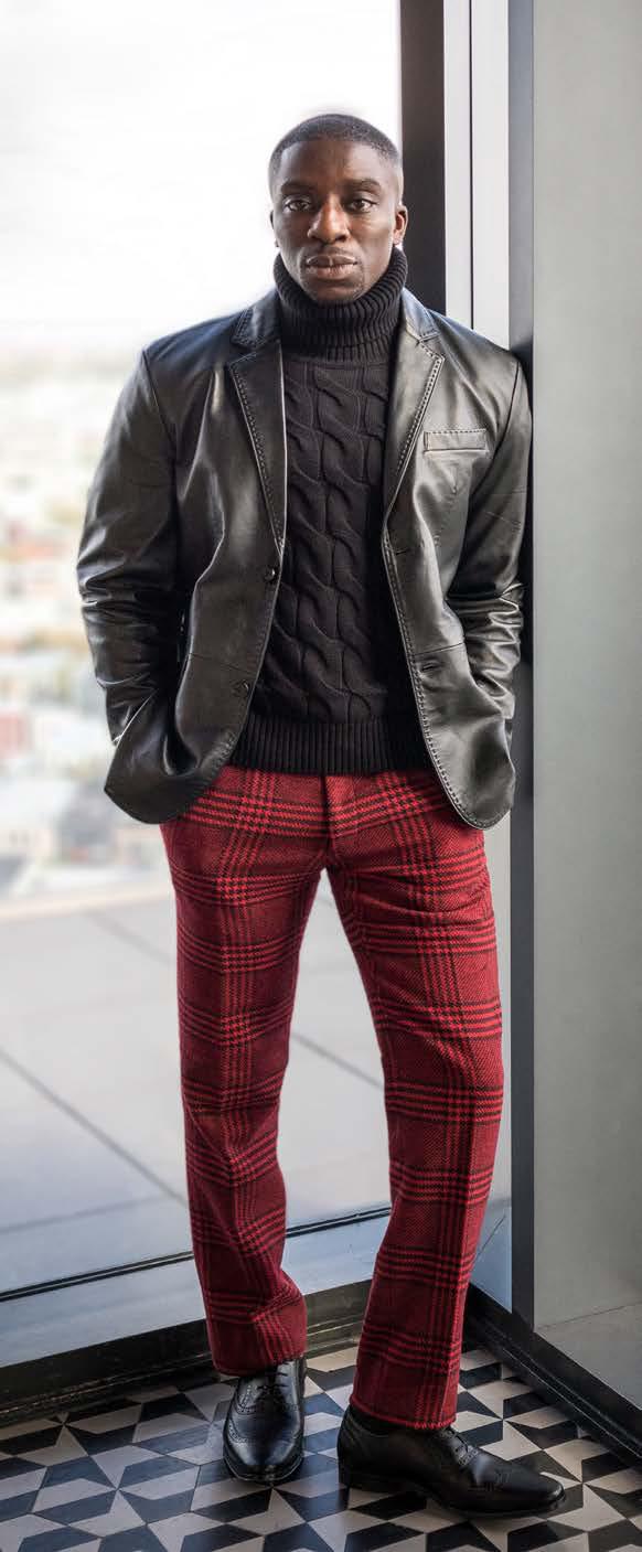 A person wearing the Inserch Lamb Leather Blazer 600-01 in black, featuring subtle pick-stitching, over a black turtleneck sweater and red plaid pants, stands indoors.