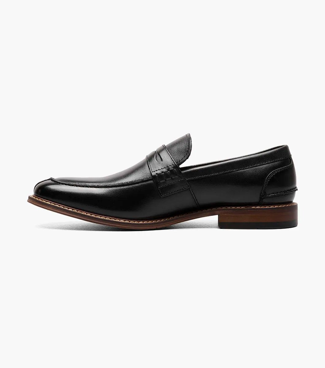 The Stacy Adams MARLOWE Algonquin Moc Toe Penny Slip On in black, featuring a refined leather upper and a contrasting brown sole, is displayed against a plain background.