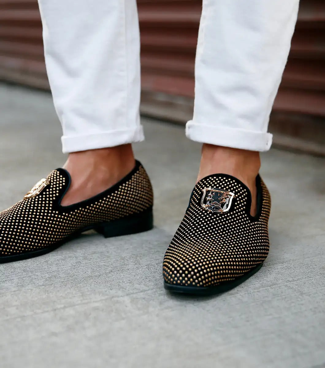 The Stacy Adams - SWAGGER Studded Slip On - Black and Gold - 25228-715 is a black loafer adorned with gold studs and a decorative emblem on top, featuring a comfortable memory foam insole.