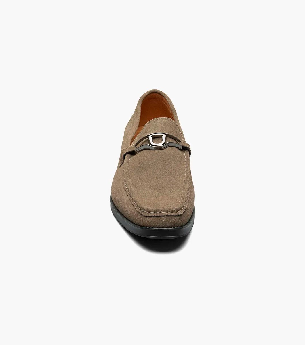 The Stacy Adams PALLADIAN Moc Toe Slip On in Fossil Suede features a brown suede leather design with a black sole, adorned with a silver buckle detail on top, and enhanced with memory foam for ultimate comfort.