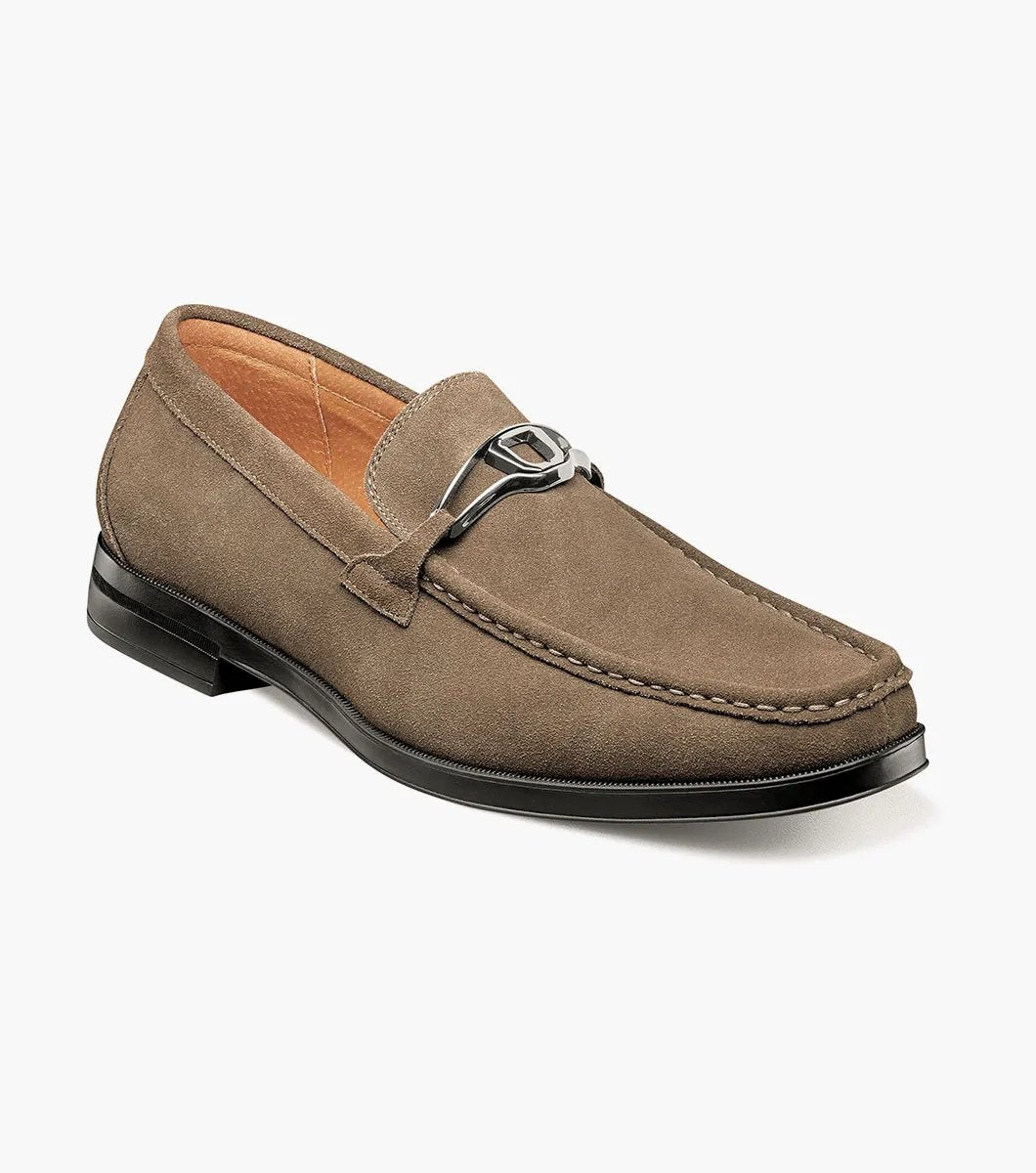 The Stacy Adams PALLADIAN Moc Toe Slip On in Fossil Suede features a brown suede leather design with a black sole, adorned with a silver buckle detail on top, and enhanced with memory foam for ultimate comfort.