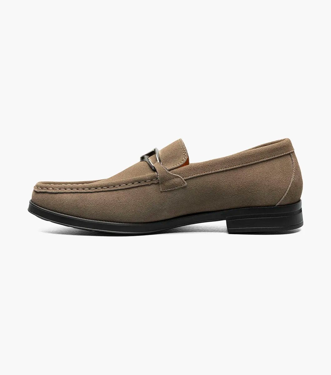 The Stacy Adams PALLADIAN Moc Toe Slip On in Fossil Suede features a brown suede leather design with a black sole, adorned with a silver buckle detail on top, and enhanced with memory foam for ultimate comfort.