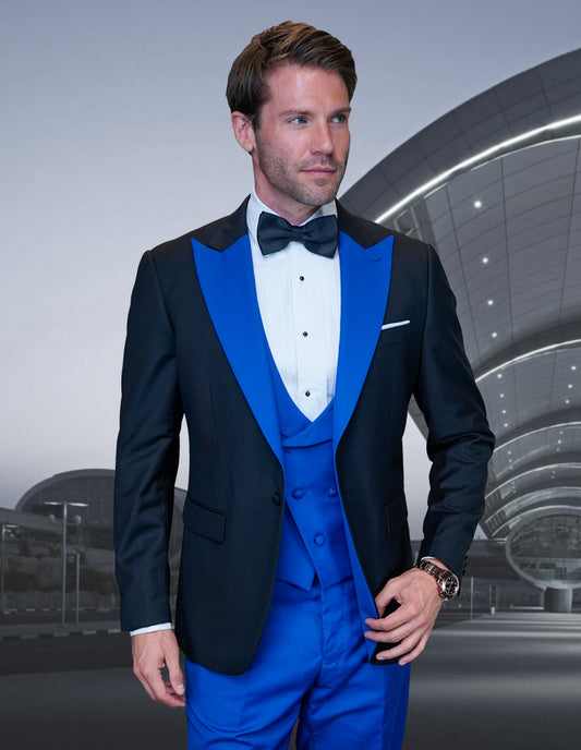 A man wearing the ARYA-ROYAL tuxedo suit from Statement Clothing, crafted from fine Italian fabric and featuring black and blue hues, poses with elegance in front of a modern architectural background.