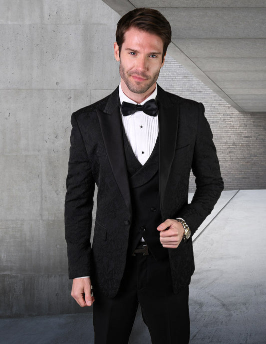 A man dressed in the STATEMENT CLOTHING | BELAGIO15-BLK by Statement Clothing, featuring a black tuxedo with a matching bow tie and tailored fit flat front pants, stands in an industrial-style hallway, looking at the camera.