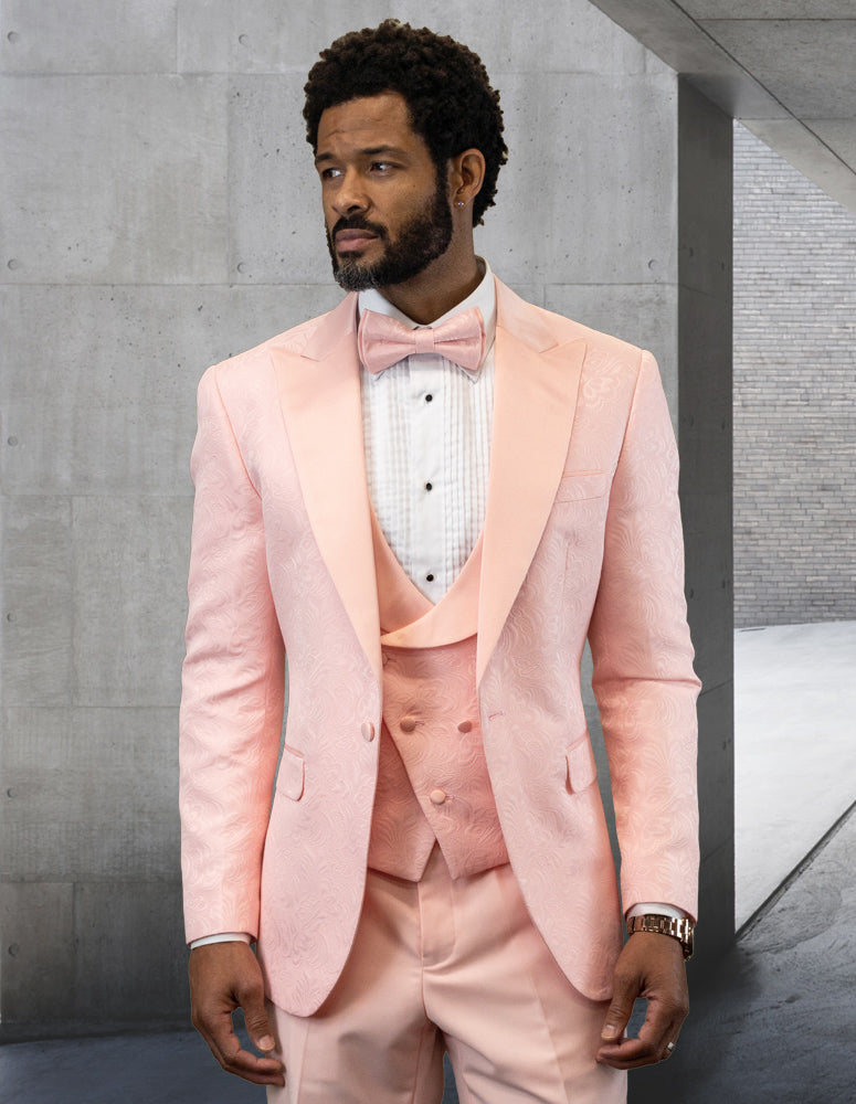 A man wearing the STATEMENT CLOTHING | BELAGIO15-BLUSH by Statement Clothing, a tailored fit, textured pink tuxedo with blush accents and a matching bow tie, stands in front of a concrete wall, looking to the side.