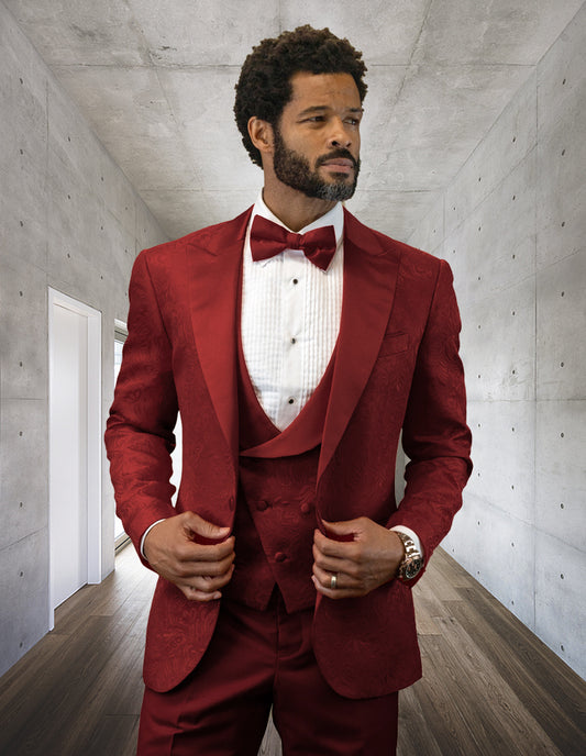 A man dressed in the BELAGIO15-BURGUNDY suit by Statement Clothing, complete with a bow tie, stands confidently in a modern room featuring concrete walls and wooden floors.