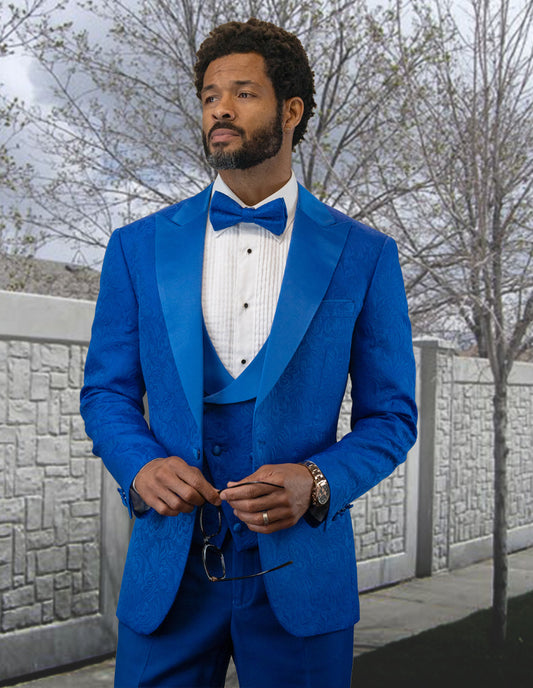 A man in a striking BELAGIO15-ROYAL tailored fit blue suit and bow tie from Statement Clothing stands regally before a stone wall and trees, holding glasses in one hand.