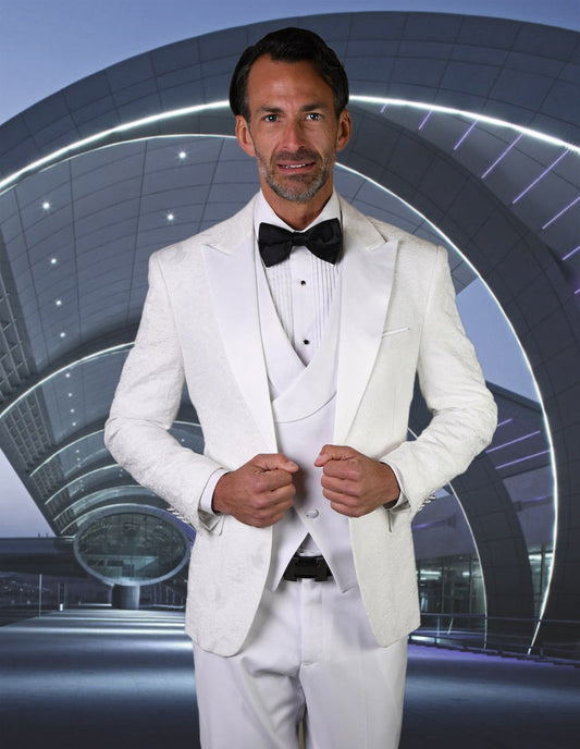 A man in a Statement Clothing | BELAGIO15-WHITE tailored-fit white tuxedo with a black bow tie stands confidently in front of a modern architectural structure at night.