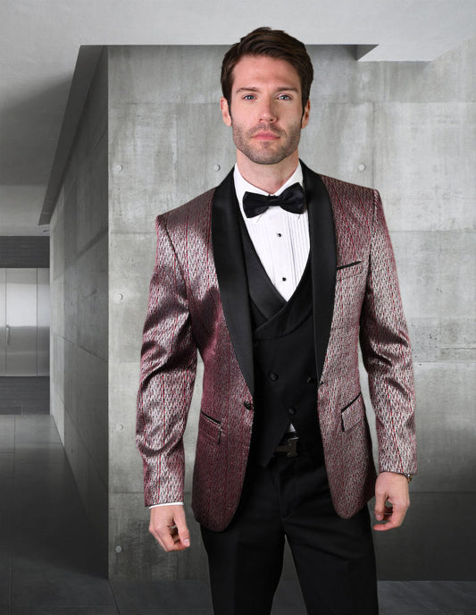 A man dressed in the STATEMENT CLOTHING | BELAGIO16-BURG tuxedo by Statement Clothing, featuring a patterned burgundy jacket with a black lapel, white shirt, and matching bow tie, stands in a modern room with concrete walls. His ensemble is completed with tailored fit flat front pants that add an elegant touch to his sophisticated appearance.