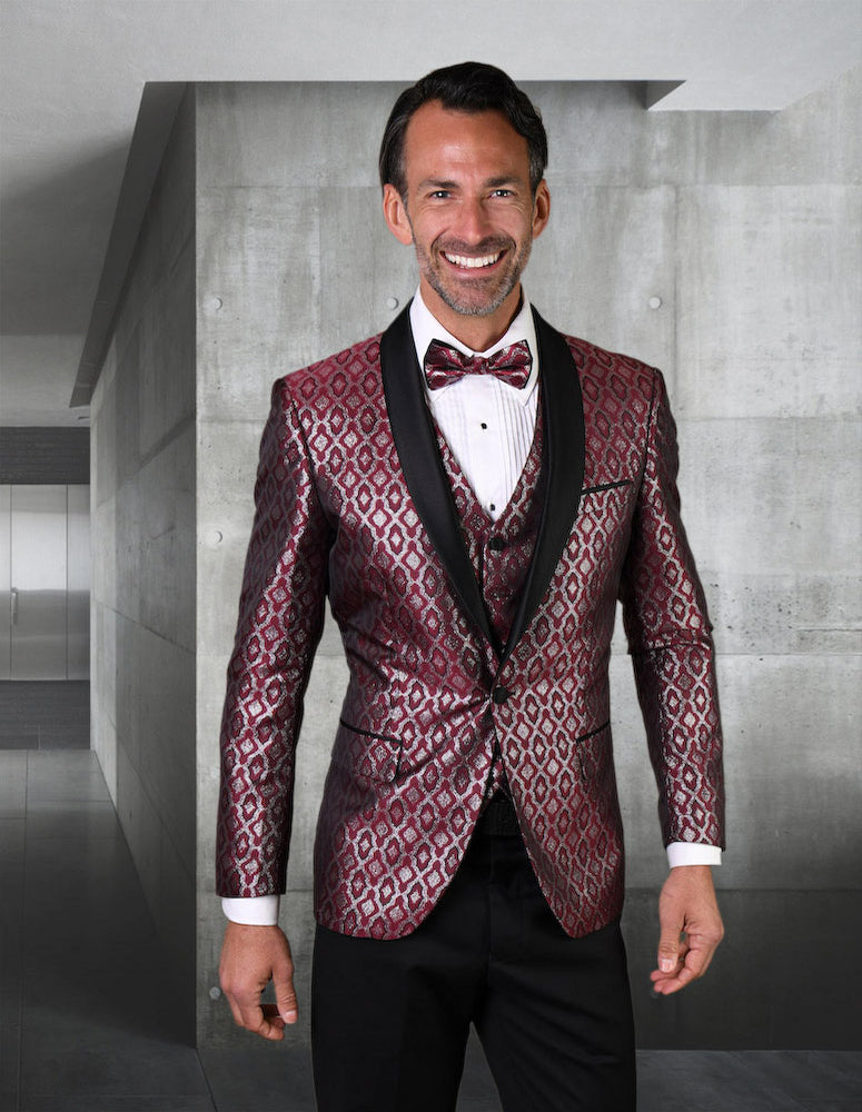 A man in a sleek, STATEMENT CLOTHING | BELAGIO21-BURGUNDY tuxedo complete with a bow tie stands smiling in a modern, minimalist room.