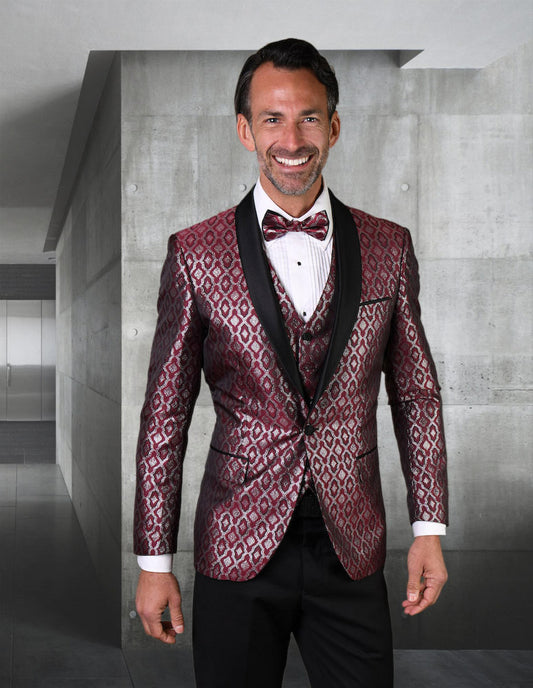 A man in a sleek, STATEMENT CLOTHING | BELAGIO21-BURGUNDY tuxedo complete with a bow tie stands smiling in a modern, minimalist room.