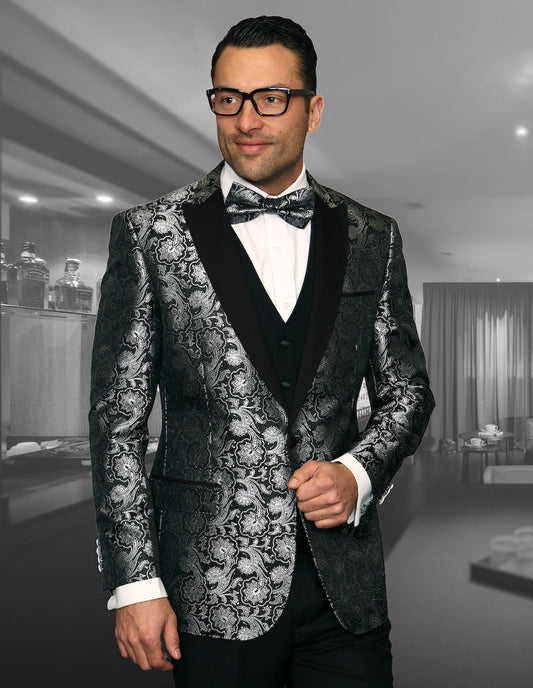 A person wearing the STATEMENT CLOTHING | BELLAGIO-4-BLACK suit from Statement Clothing, featuring a black and silver pattern with slim fit flat front pants, white shirt, bow tie, and glasses stands in a room with blurred shelves and curtains in the background.