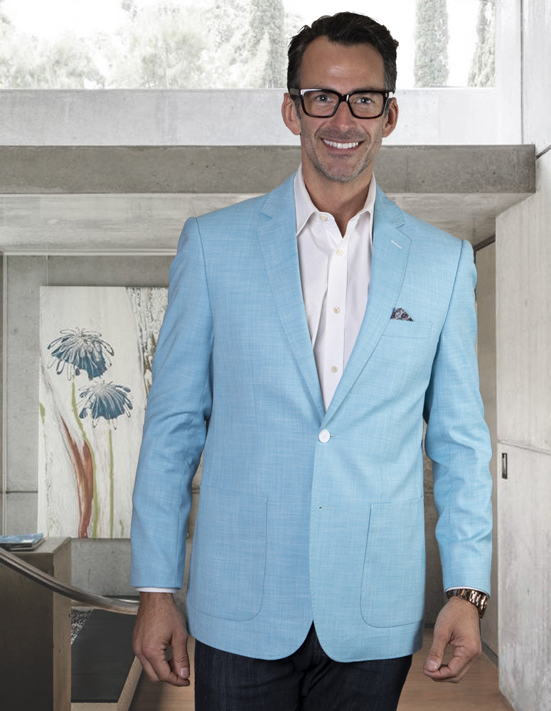 A man wearing the BELUCHI-TURQUOISE suit jacket from Statement Clothing, crafted from Super 150's Italian fabric, is indoors smiling. He also wears glasses and stands in front of modern decor, which includes a painting adorned with turquoise blue flowers.