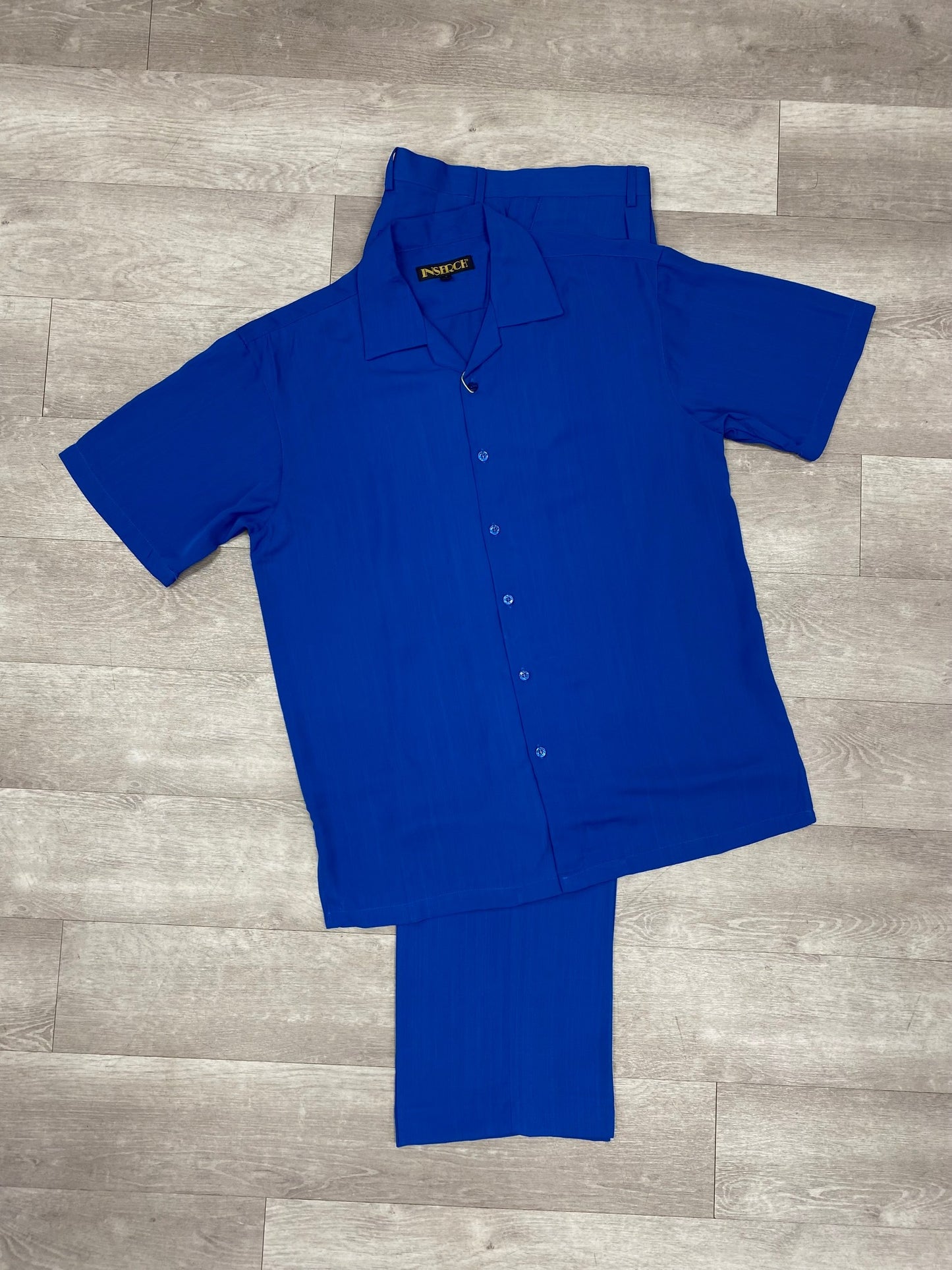 The trendy ensemble includes the Inserch 2pc 9356 Imperial Blue, consisting of a short-sleeve button-up microfiber shirt and matching pants in a striking blue shade, all elegantly displayed on a wooden floor. Perfect for any relaxed occasion, this outfit by Inserch offers both style and comfort.