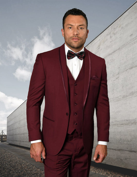 A man in the STATERMENT CLOTHING | CAESAR-BURGUNDY suit by Statement Clothing, paired with a bow tie, stands against a concrete wall backdrop.