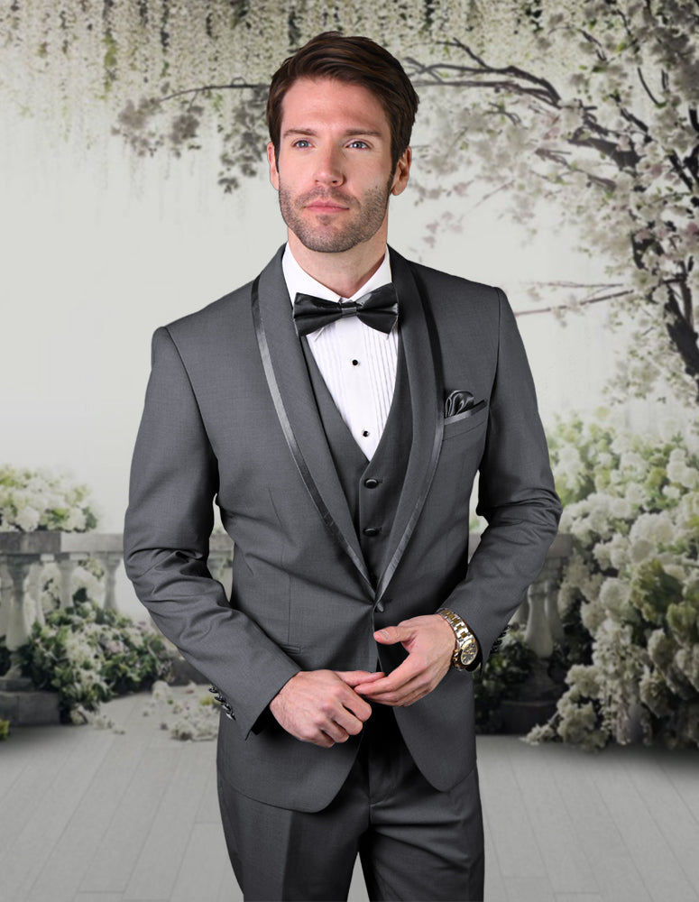 A man dressed in the STATEMENT CLOTHING | CAESAR-CHARCOAL tuxedo from Statement Clothing, paired with a crisp white shirt and an elegant bow tie, stands against a floral backdrop.