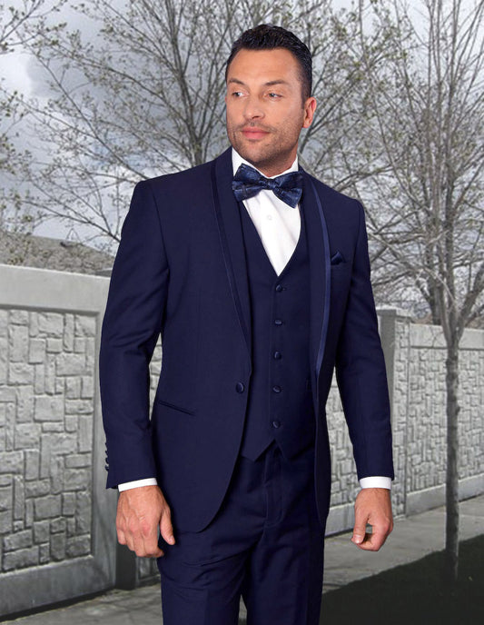 A man wearing the CAESAR-NAVY formal suit by Statement Clothing, featuring a tailored fit and complemented with a bow tie, stands outdoors in front of a stone wall and bare trees.