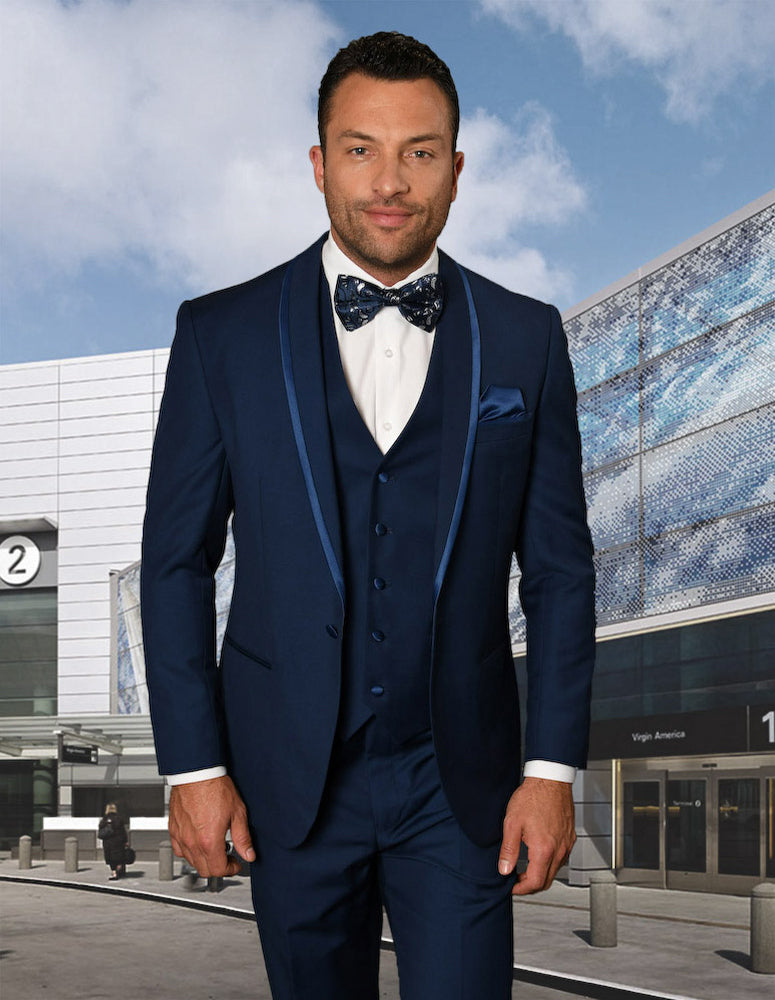 Wearing the STATEMENT CLOTHING | CAESAR-SAPPHIRE by Statement Clothing, a man in a tailored fit suit and bow tie stands in front of a modern building with a glass facade under a partly cloudy sky.