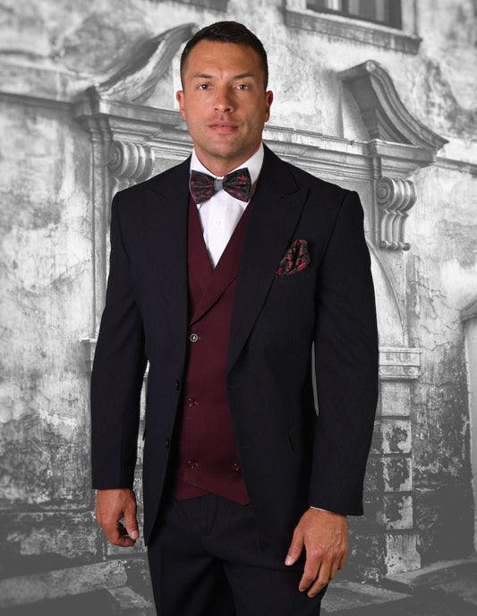 A man in a STATEMENT CLOTHING | CAMDEN-BLACK suit featuring a striking Italian wool texture and complemented by a burgundy vest stands confidently against a textured, grey wall background.