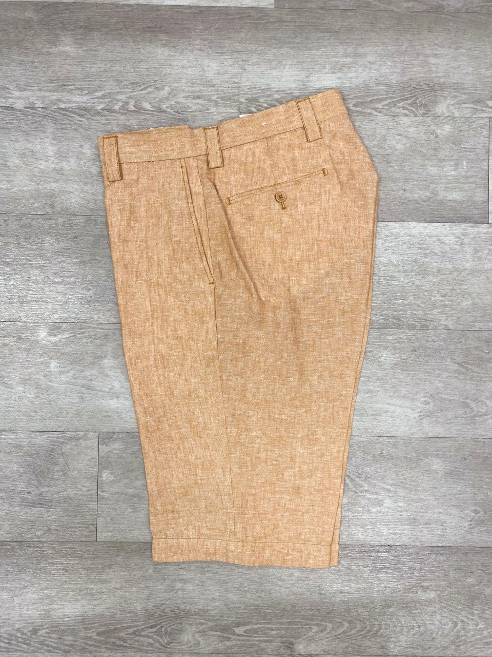 The Inserch Premium Linen Flat Front Shorts in camel, crafted from Belgian Flax, featuring button and pocket details, are elegantly displayed on a wooden surface. Perfect for enhancing your summer wardrobe.