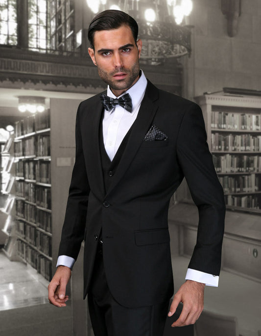 A man in a CORTA-BLACK ultra slim fit tuxedo from Statement Clothing, complete with a bow tie, stands in an elegant library styled with vintage charm, tall shelves, and chandeliers, radiating sophistication and style.