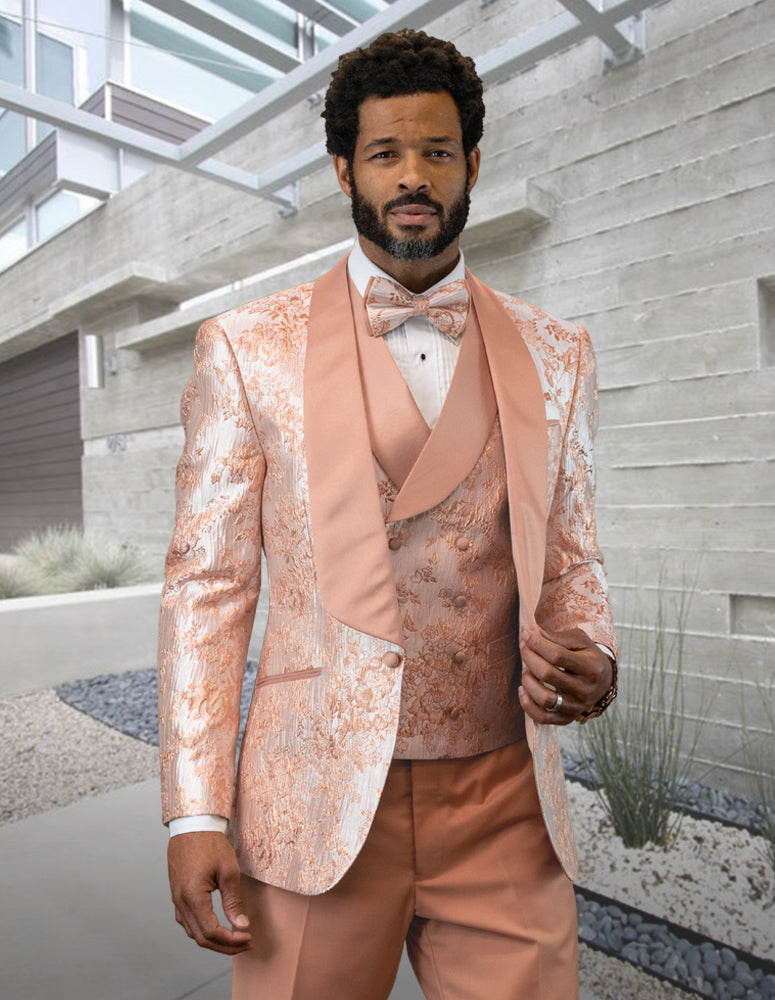 A man dressed in the DELANO-BLUSH tuxedo by Statement Clothing, featuring a peach and white floral shawl lapel and a bow tie, stands in a modern architectural setting.