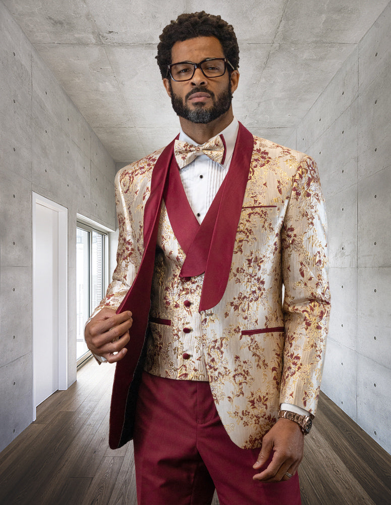 A man is wearing the STATEMENT CLOTHING | DELANO-BURGUNDY floral-patterned suit with a shawl lapel tuxedo, perfectly paired with a burgundy vest and pants from Statement Clothing, as he stands in a modern hallway.