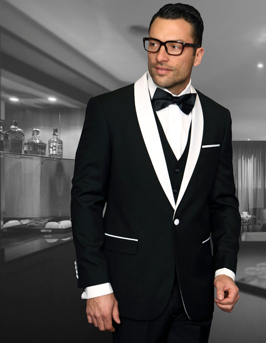 A man in the Statement Clothing's ENCORE-BLACK tuxedo, featuring a tailored fit and flat front pants, stands in an elegantly furnished room, his bow tie perfectly knotted as he looks to the side.