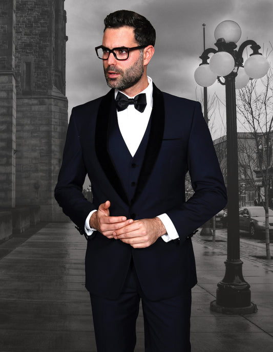 A man wearing the ENCORE-V-NAVY-1 ensemble by Statement Clothing, complete with a sleek jacket featuring a velvet lapel and matching accessories, stands on a city street with a historical building and vintage streetlamp adorning the background.