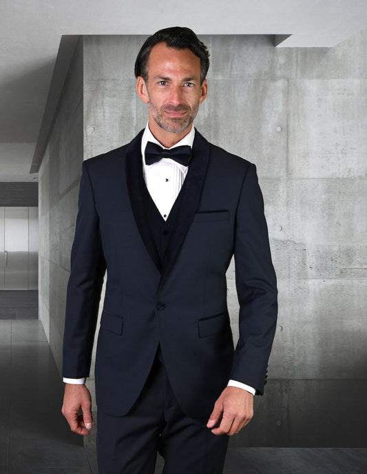 A man wearing the ENCORE-V-NAVY tuxedo by Statement Clothing, featuring a tailored fit with a navy velvet lapel and bow tie, stands indoors against a concrete wall background.