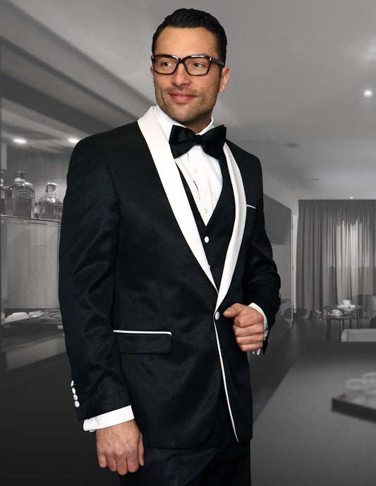 A person wearing the STATEMENT CLOTHING | ENZO-6-BLACK, a formal black shawl lapel tuxedo from Statement Clothing, stands in a dimly lit room with shelves and a curtain in the background while sporting glasses.