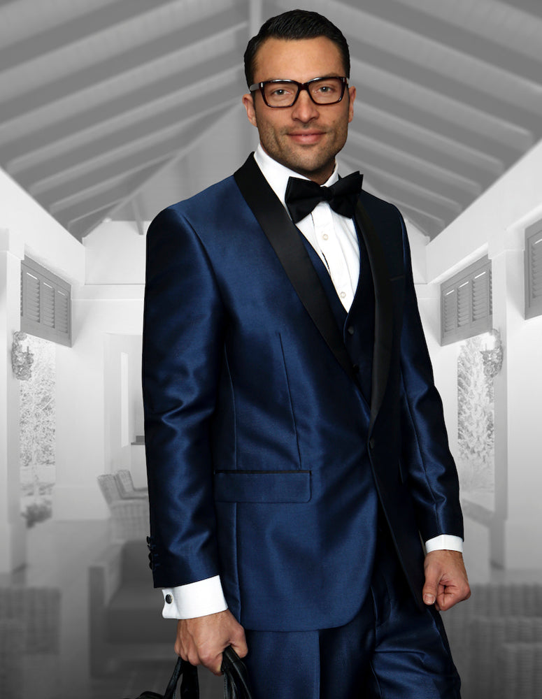 A man wearing a tailored-fit Statement Clothing ENZO-7-INDIGO tuxedo, complete with a black bow tie and glasses, stands indoors holding a briefcase.