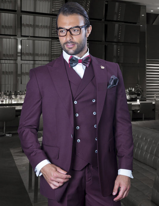 A man dressed in the Florence-Burgundy suit from Statement Clothing, featuring wide-leg pants and a bow tie, stands in a modern lounge, with the understated elegance of Italian wool elevating his presence as the bar softly gleams in the background.