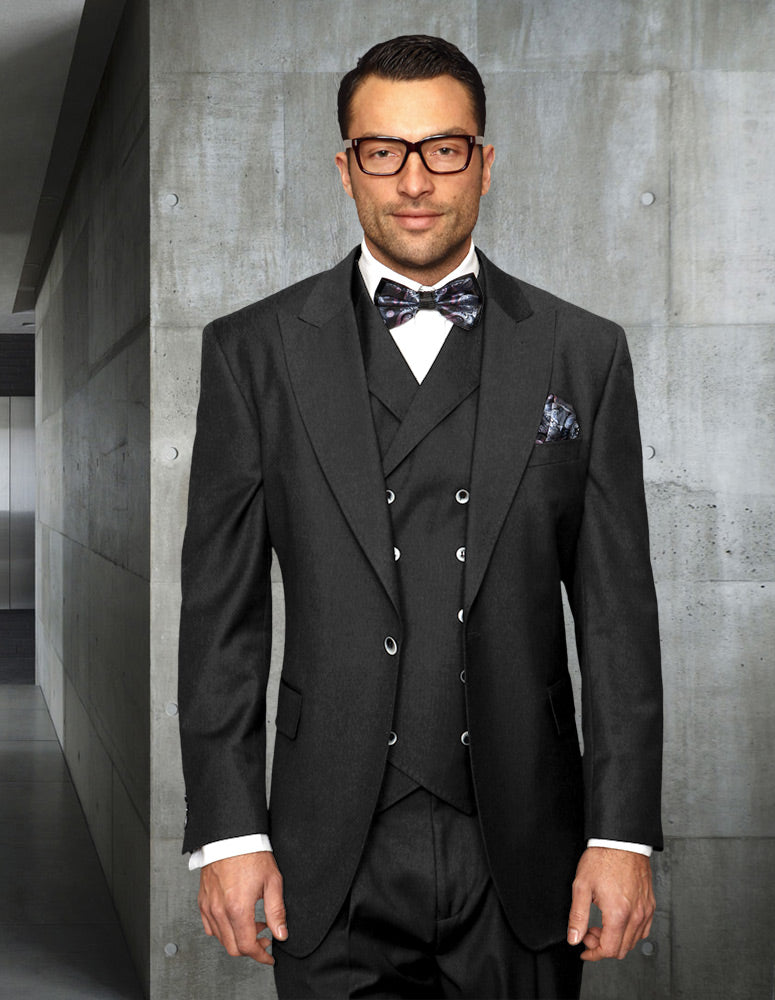 A man in the STATEMENT CLOTHING | FLORENCE-CHARCOAL three-piece suit by Statement Clothing, complete with a bow tie and glasses, stands in a modern, industrial-style hallway.