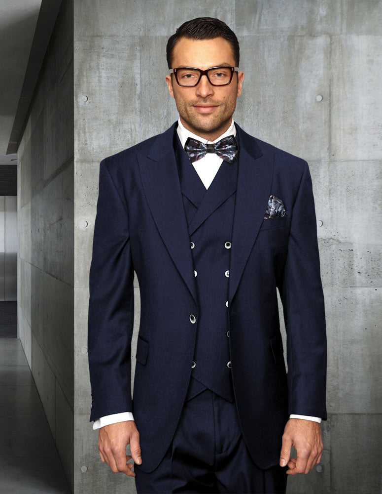 A man wearing the STATEMENT CLOTHING | FLORENCE-NAVY by Statement Clothing, complete with a navy Italian wool three-piece suit and bow tie, stands in a modern, industrial-style hallway.