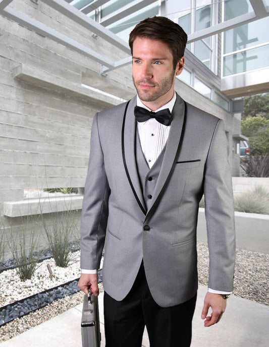 A man dressed in the GENOVA-GREY tuxedo by Statement Clothing, featuring flat front pants, and adorned with a black bow tie, stands in a sleek modern architectural environment of concrete and glass while holding a briefcase.