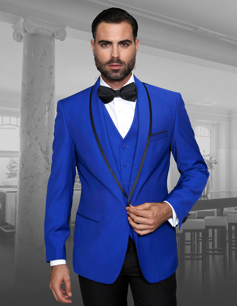 A man wearing the GENOVA-ROYAL tuxedo by Statement Clothing stands confidently, its royal blue hue with black trim and perfect tailored fit creating a striking presence, while his bow tie adds an elegant touch in a room adorned with large windows and a marble pillar.