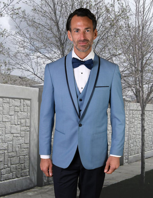 A man in a STATEMENT CLOTHING | GENOVA-STEELBLUE tuxedo from Statement Clothing, featuring a tailored fit in steel blue with a black lapel and bow tie, stands in front of a stone wall and trees.