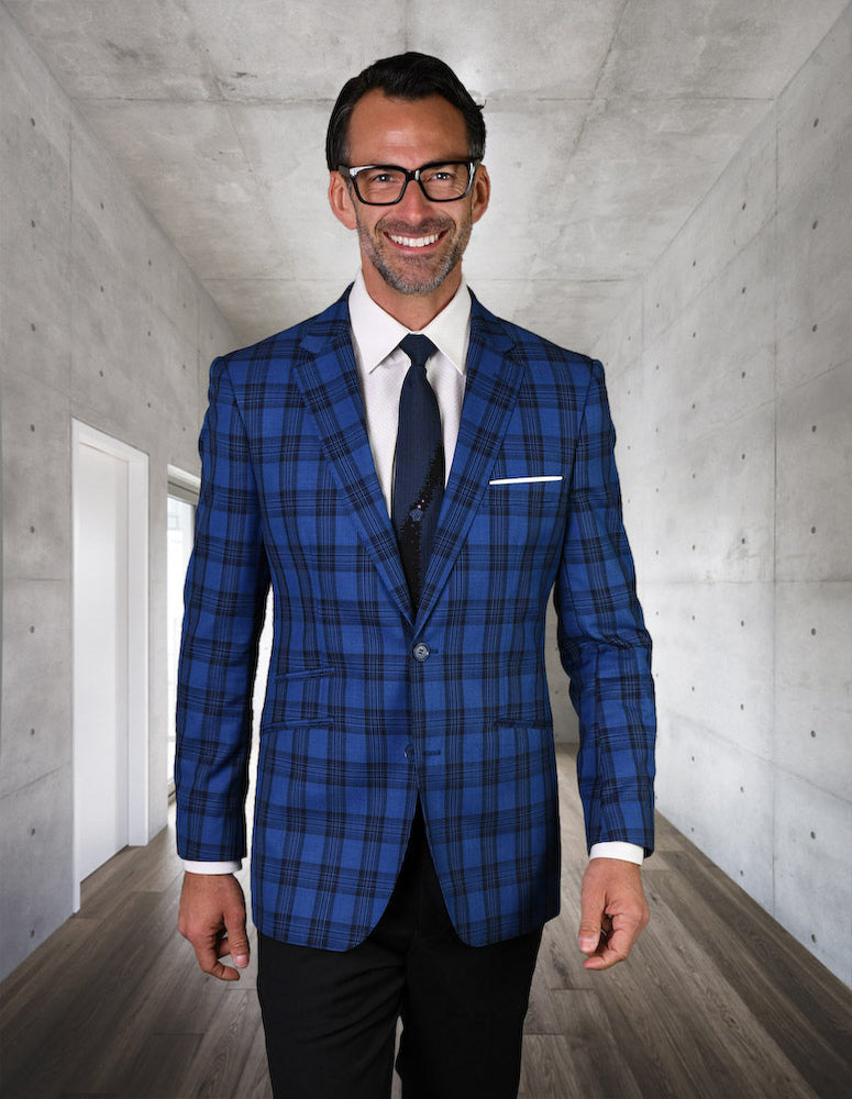 A man wearing a tailored fit sapphire plaid suit and glasses stands in a modern hallway, smiling. The jacket he's wearing is the GIGLIO-12-SAPPHIRE by Statement Clothing, made from Super 150's fabric for a blend of elegance and comfort.