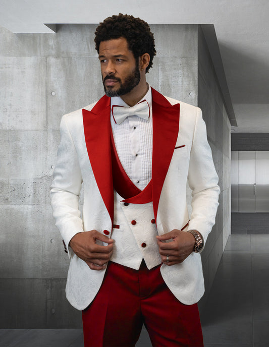 A man dressed in the modern-fit STATEMENT CLOTHING | HILTON-RED tuxedo from Statement Clothing, featuring red accents and a bow tie, stands indoors against a sleek concrete wall.