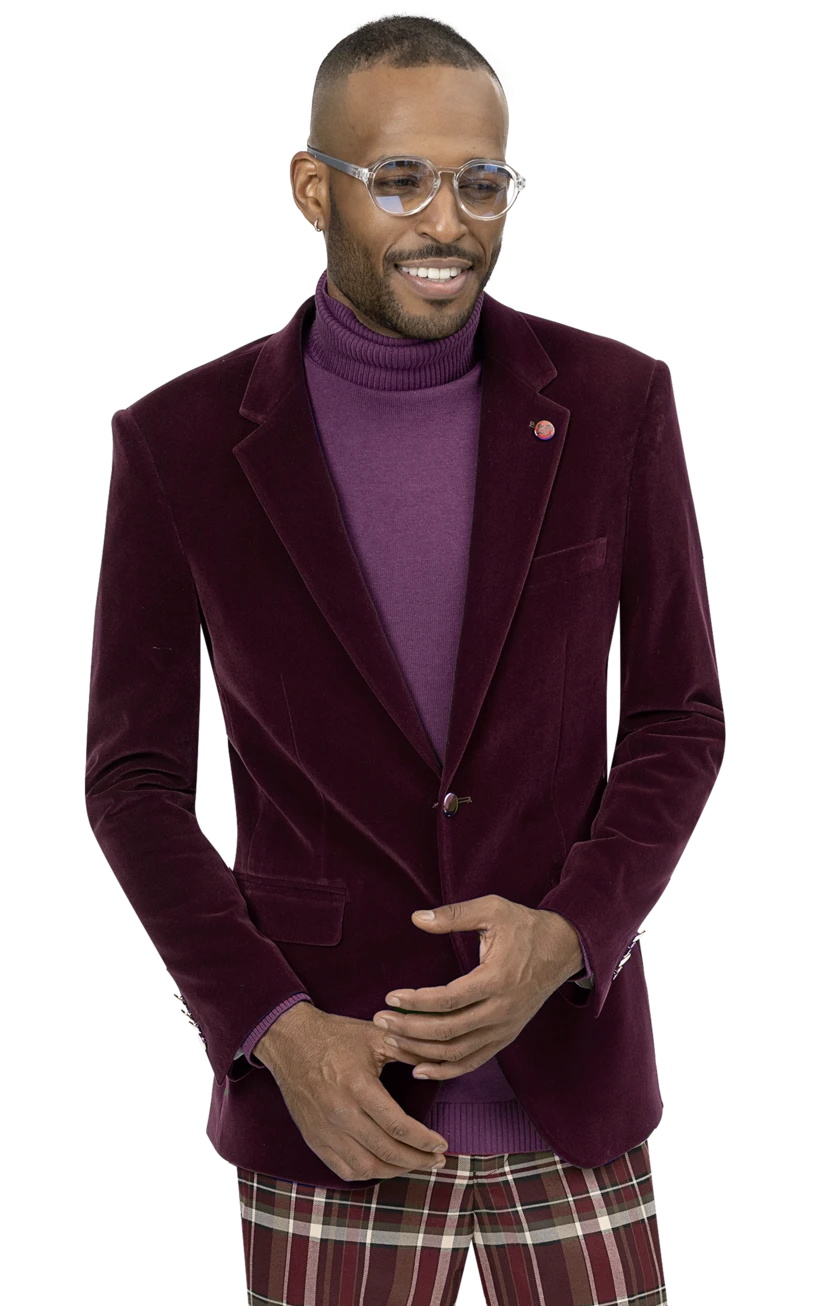 A man in an EJ Samuel Burgundy Velvet Blazer J134, featuring gold buttons, styled with a purple turtleneck and clear glasses, completes his look with plaid pants as he smiles and poses with hands together.