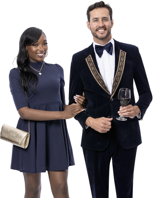 A woman in a dress embellished with sequins stands beside a man dressed in an EJ Samuel Navy Suit JP110, complete with a bow tie. Both are smiling as the man holds a glass of wine and the woman carries a gold clutch.