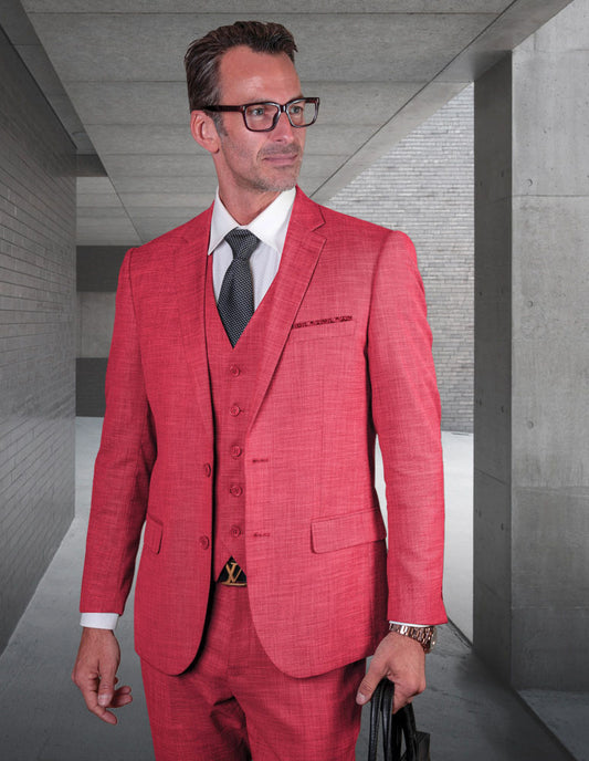 A person in a slim fit magenta suit from Statement Clothing's LAZARO-MAGENTA collection, crafted from fine Italian fabric and wearing glasses, stands in a modern, concrete hallway, clutching a black bag.