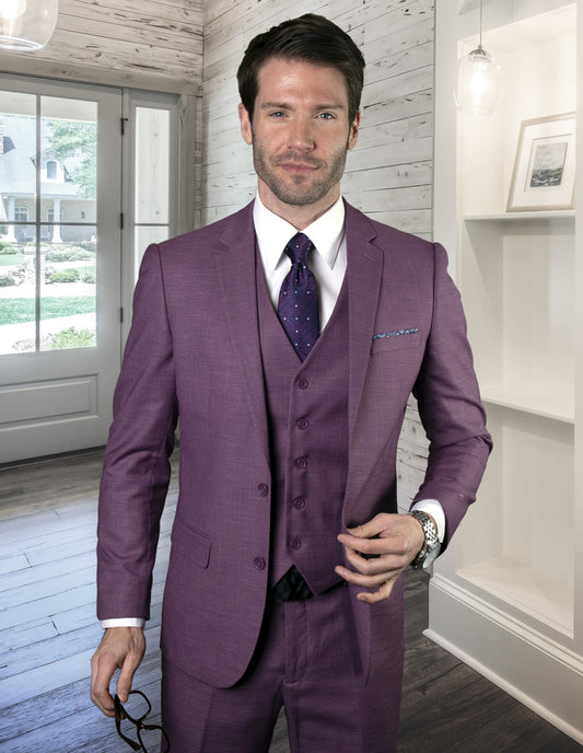 A man wearing the LAZARO-MAUVE suit from Statement Clothing, tailored in a slim fit from Italian fabric, holds eyeglasses in one hand while standing in an elegant room featuring white wood paneling and abundant natural light.