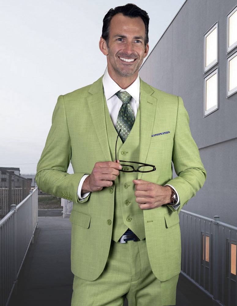 A man wearing the STATEMENT CLOTHING | LAZARO-PISTACHIO suit from Statement Clothing, crafted from luxurious Italian fabric, holds glasses while standing on a modern building walkway.