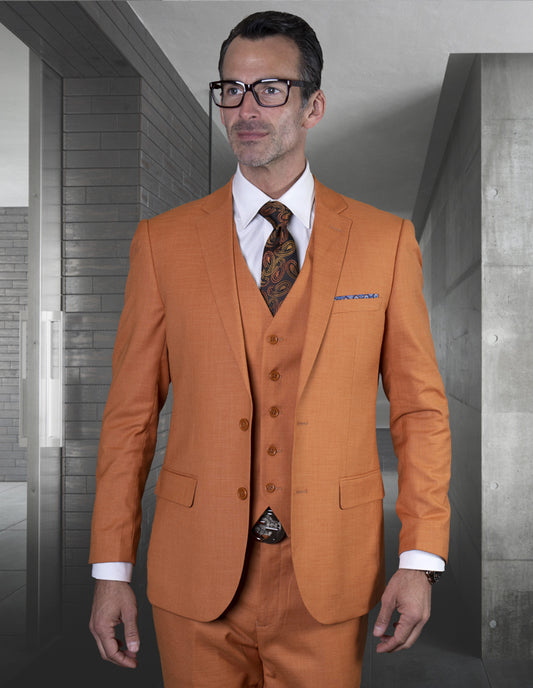 A person wearing the LAZARO-RUST slim fit three-piece suit from Statement Clothing, crafted from luxurious Italian fabric, exudes elegance against a backdrop of sleek grey walls in a modern setting. The ensemble is completed with a white shirt and patterned tie.