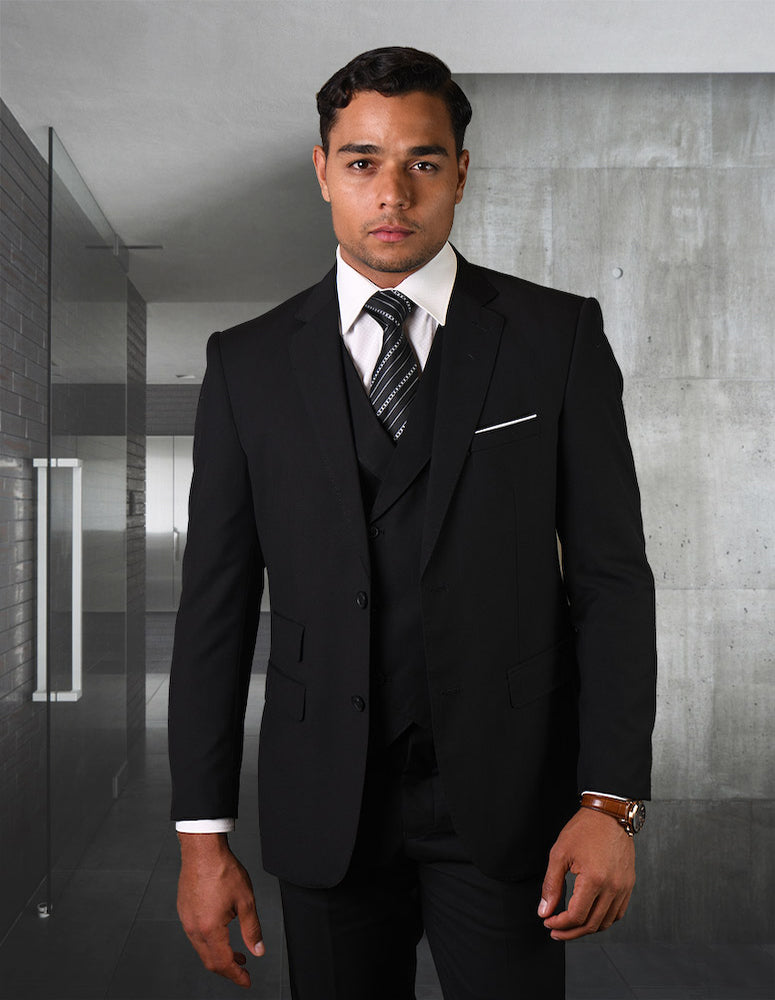 A man wearing the STATEMENT CLOTHING | LAZIO-BLACK suit from Statement Clothing stands in a modern building hallway, exuding sophistication. The Super 150's fabric and flat front pants create a tailored fit that enhances his elegant silhouette against the backdrop of glass walls and concrete detailing.