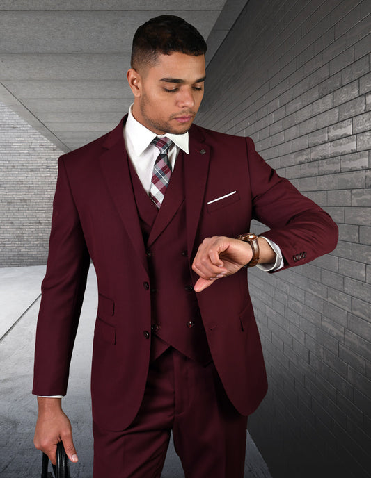 Dressed in a Lazio-Burgundy suit by Statement Clothing, a man with flat front pants checks his wristwatch while holding a briefcase. He stands in a grey, modern hallway.